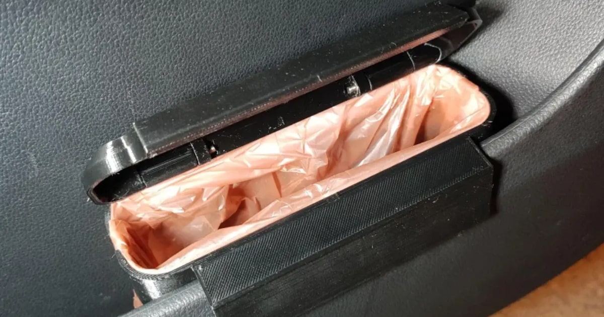Primer plano de un pequeño cubo de basura negro impreso en 3D, colocado en el hueco de la puerta de un coche. El cubo tiene una tapa abatible abierta y una bolsa de plástico en su interior, sujeta por un aro diseñado para mantenerla en su sitio